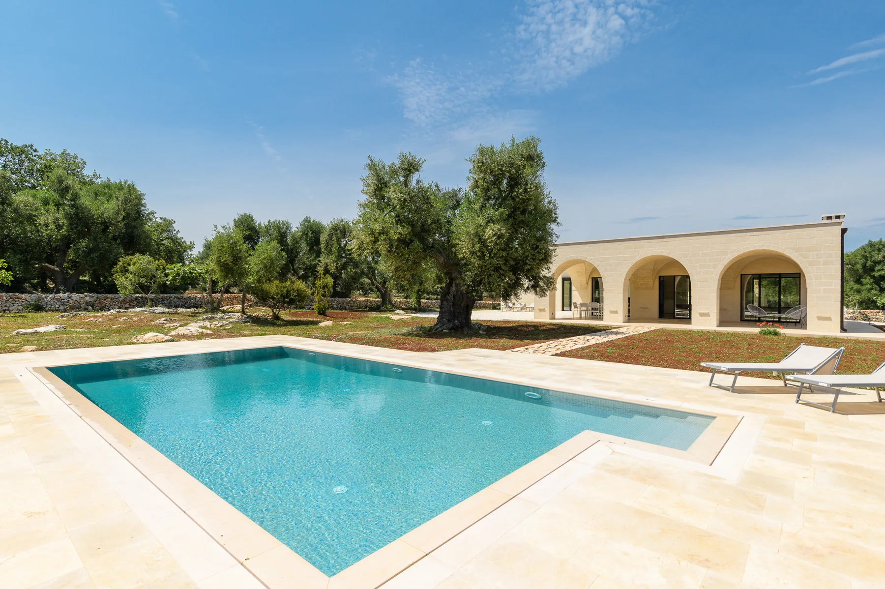 An outdoor swimming pool sits beside a low building with arched openings and trees.