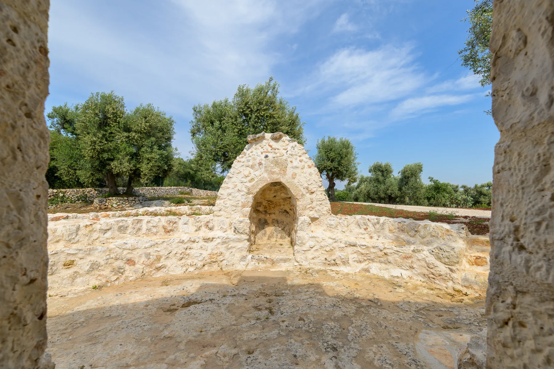 A stone courtyard with a small arched alcove and trees beyond the wall.