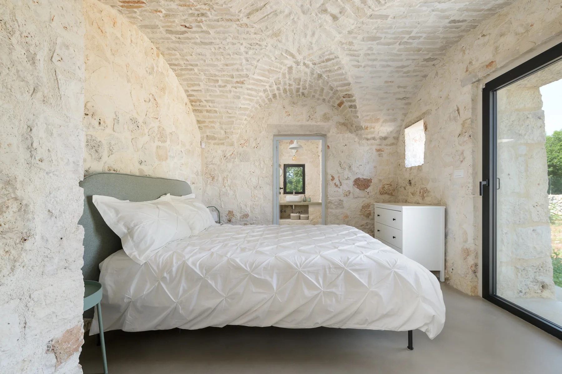 A bedroom with a bed, pale stone walls, and a glass door.