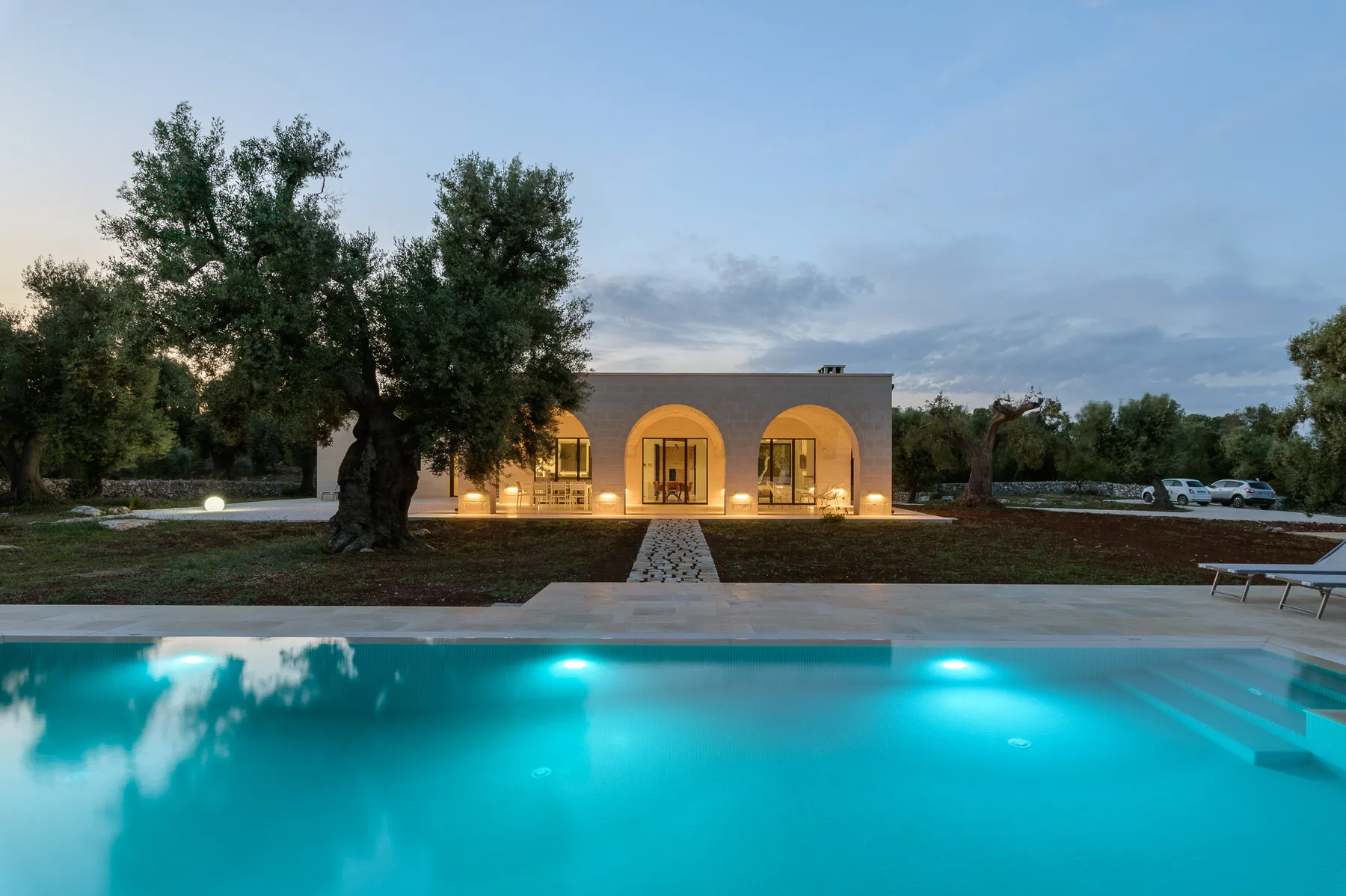 A lit building stands beyond a swimming pool, with trees and a walkway in view.