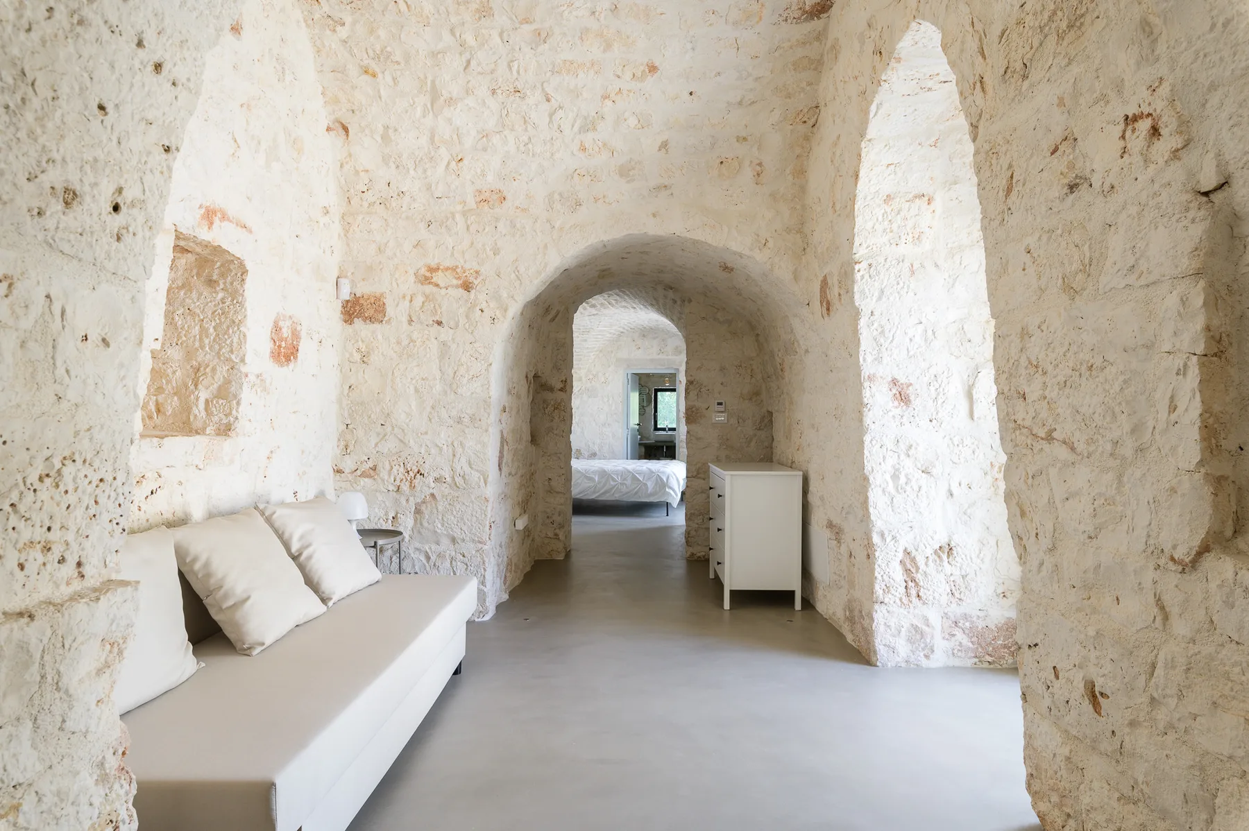 A pale stone room has a white sofa and an arched passage leading to a bedroom.