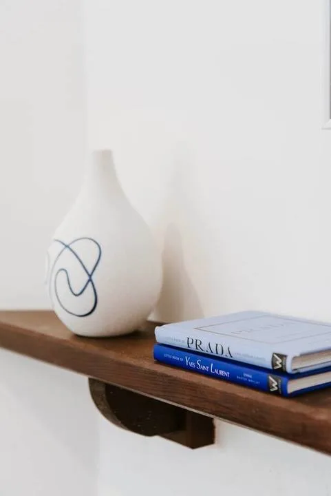 Un jarrón decorativo y unos libros están sobre una repisa de madera contra una pared blanca.