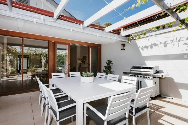 Una zona exterior cubierta para comer, con una mesa blanca larga junto a una parrilla.
