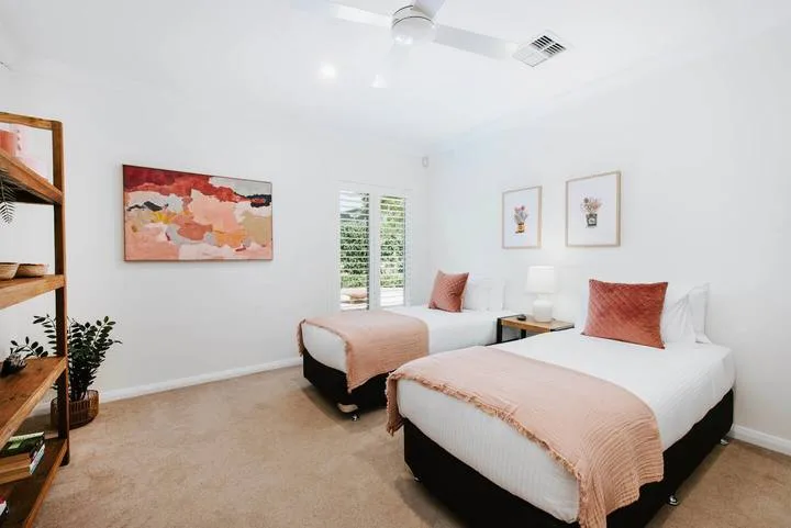 Un dormitorio luminoso con dos camas vestidas con ropa de cama blanca y color durazno.