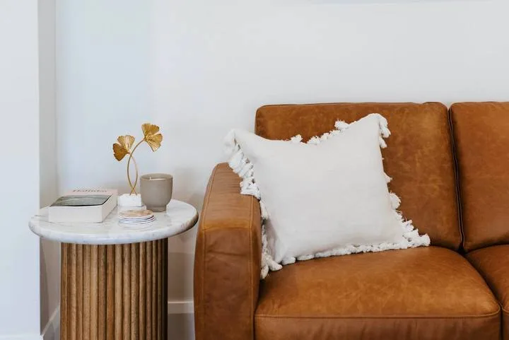 Un sofá color caramelo está junto a una mesa auxiliar redonda con libros y un jarrón.
