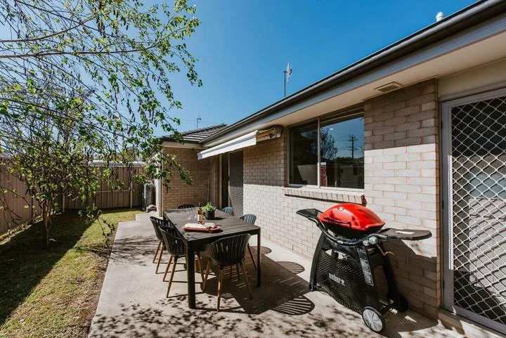 Comedor exterior pavimentado junto a una casa de ladrillo, con una mesa y una barbacoa.