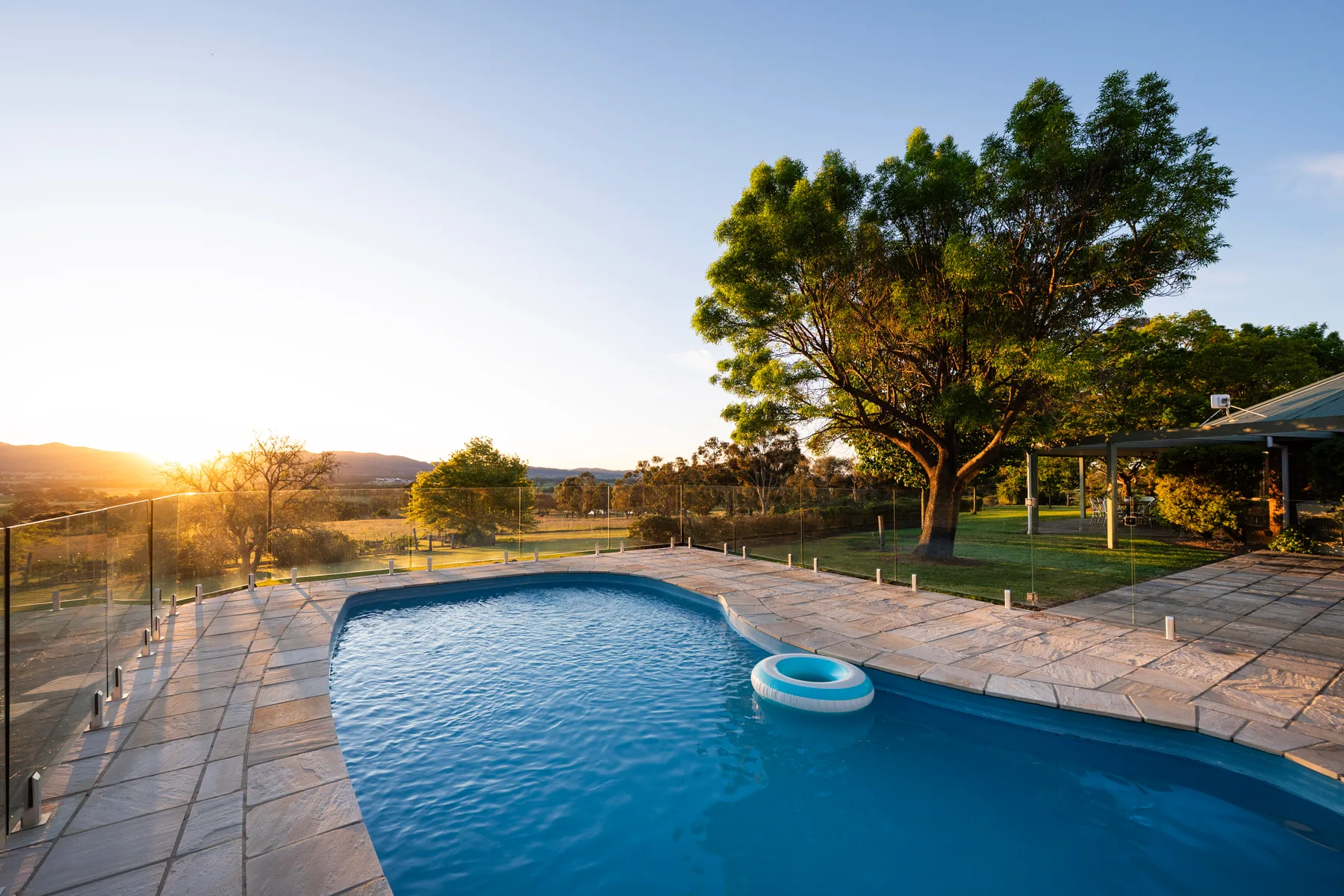An outdoor swimming pool with a small inflatable ring sits beside a landscaped yard at sunset.