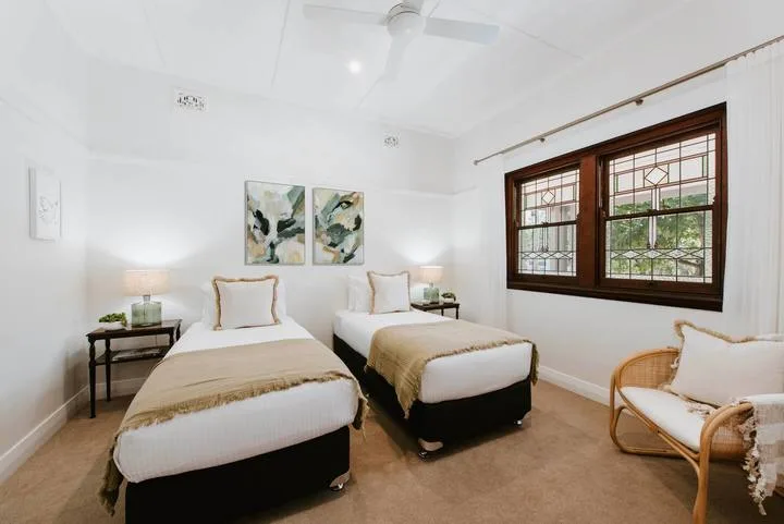 Un dormitorio luminoso con dos camas, cuadros y una ventana grande.