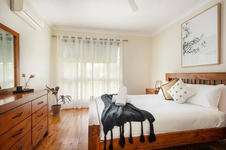 Un dormitorio luminoso con cama de madera, ropa de cama blanca y cortinas translúcidas.