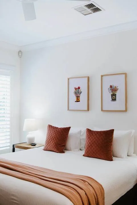 Un dormitorio con una cama bien tendida, cojines color óxido y láminas florales enmarcadas.