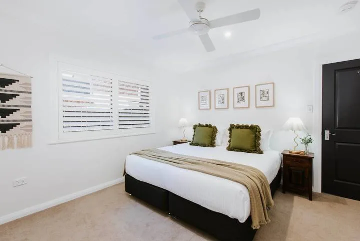 Un dormitorio luminoso con una cama, cuadros y ventanas con persianas.