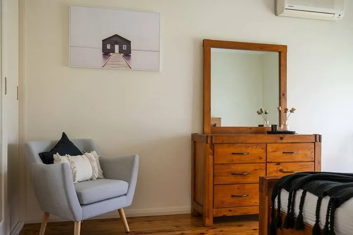 Un dormitorio con un sillón gris junto a una cómoda de madera y un espejo.