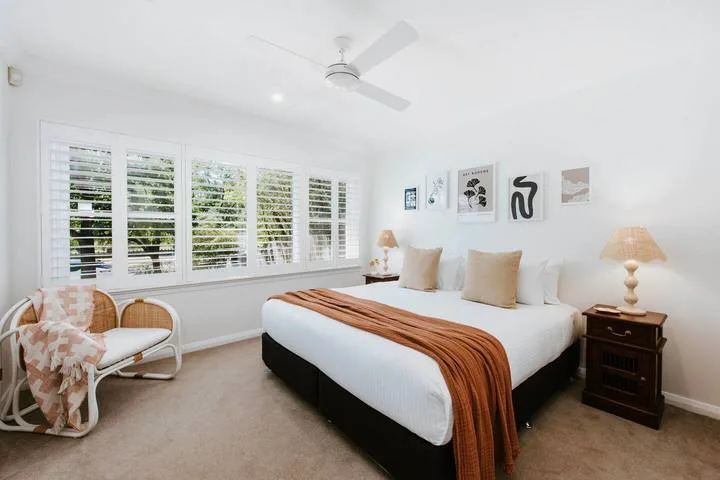 Un dormitorio luminoso con una cama arreglada y amplias ventanas con persianas.