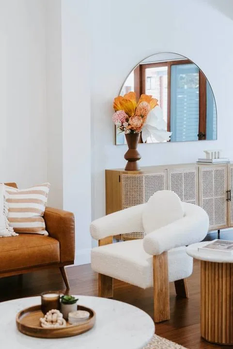 Una sala luminosa con un sillón blanco, un sofá marrón y un espejo redondo en la pared.