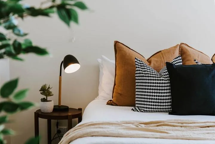 Una cama de dormitorio está arreglada con cojines estampados y una lámpara de noche.