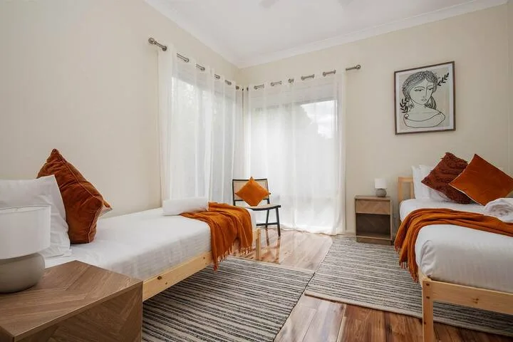 Un dormitorio luminoso con dos camas, cortinas transparentes y detalles naranjas.