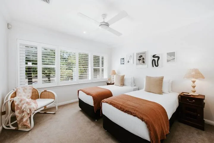 Un dormitorio luminoso con dos camas, ventanas grandes y un ventilador de techo.