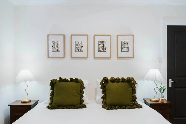 Un dormitorio con una cama blanca, almohadas verdes, mesitas y cuadros enmarcados.