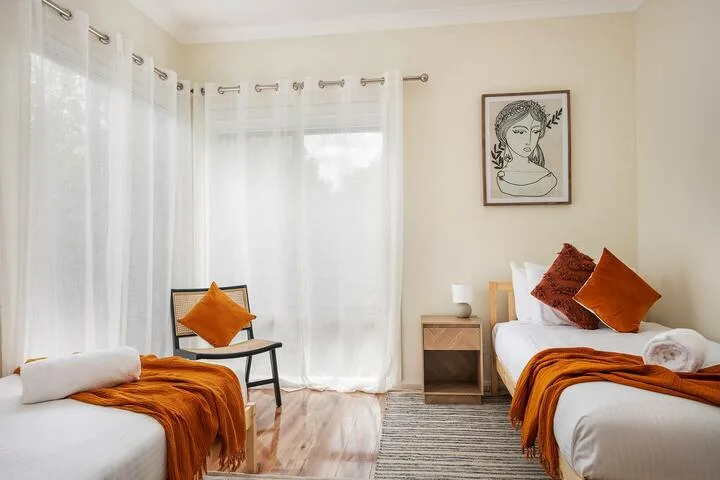 Un dormitorio luminoso con dos camas, cortinas transparentes y ropa de cama naranja.