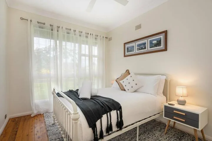 Un dormitorio luminoso con una cama blanca, una manta oscura y cortinas translúcidas.