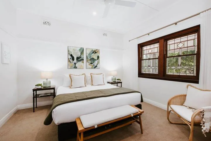 Un dormitorio luminoso con una cama, mesitas de noche y una gran ventana con marco de madera.