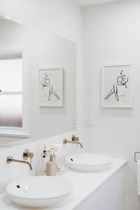 Un baño luminoso con dos lavabos blancos sobre encimera y cuadros sencillos.