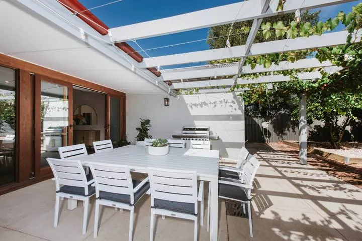 Un comedor exterior está bajo una pérgola blanca junto a puertas de vidrio.