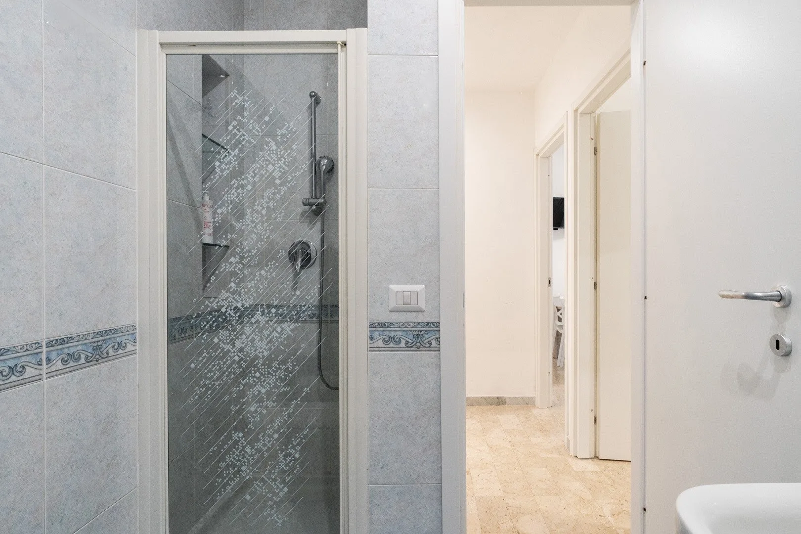 A tiled bathroom with a glass shower beside an open doorway.