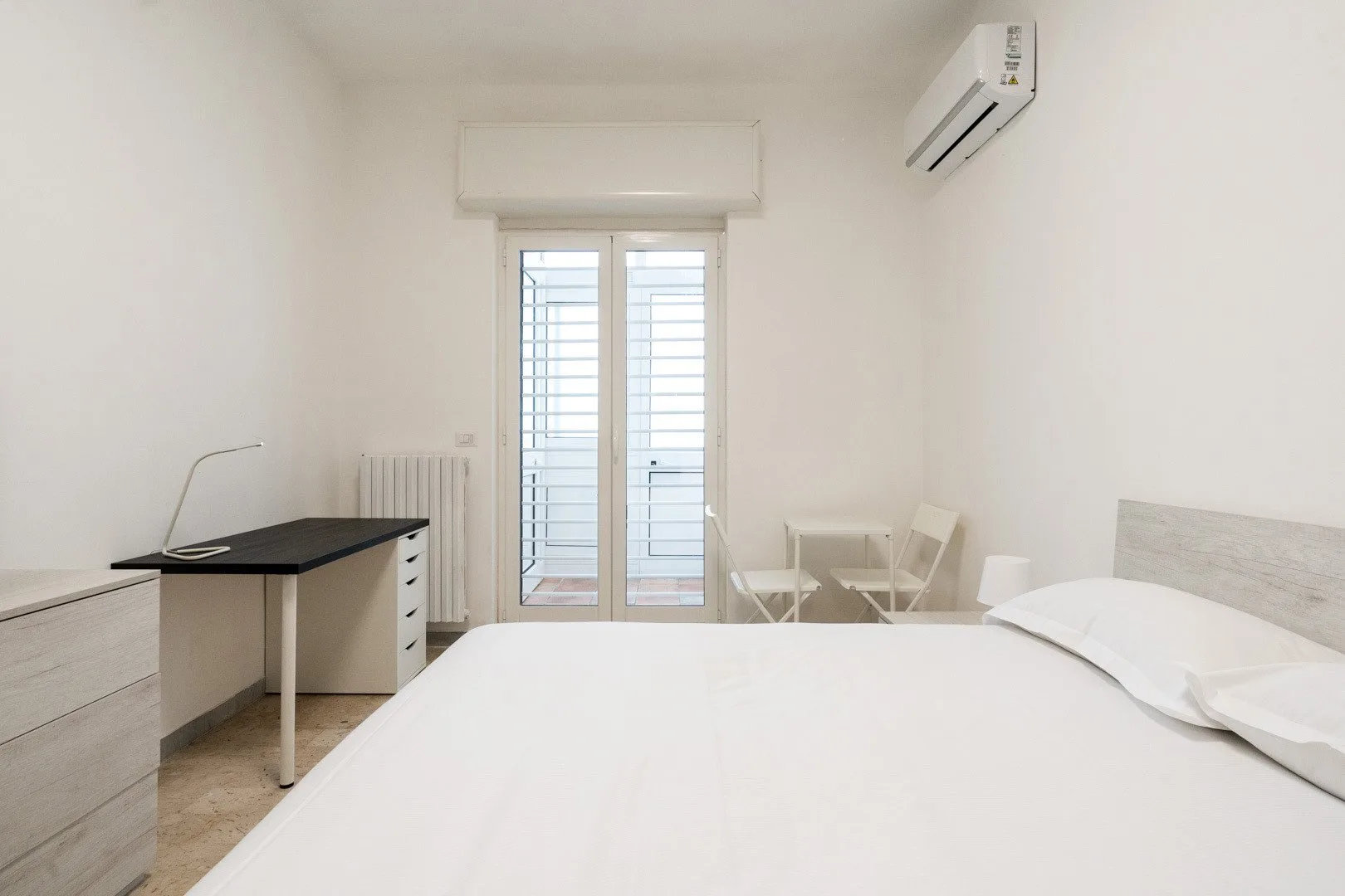 A bright bedroom with a bed, desk, and glass doors opening to a balcony.