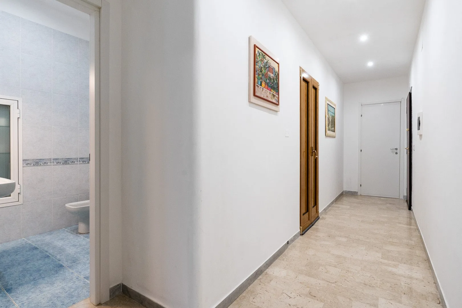 A bright hallway with framed artwork, wooden doors, and a pale tiled floor.