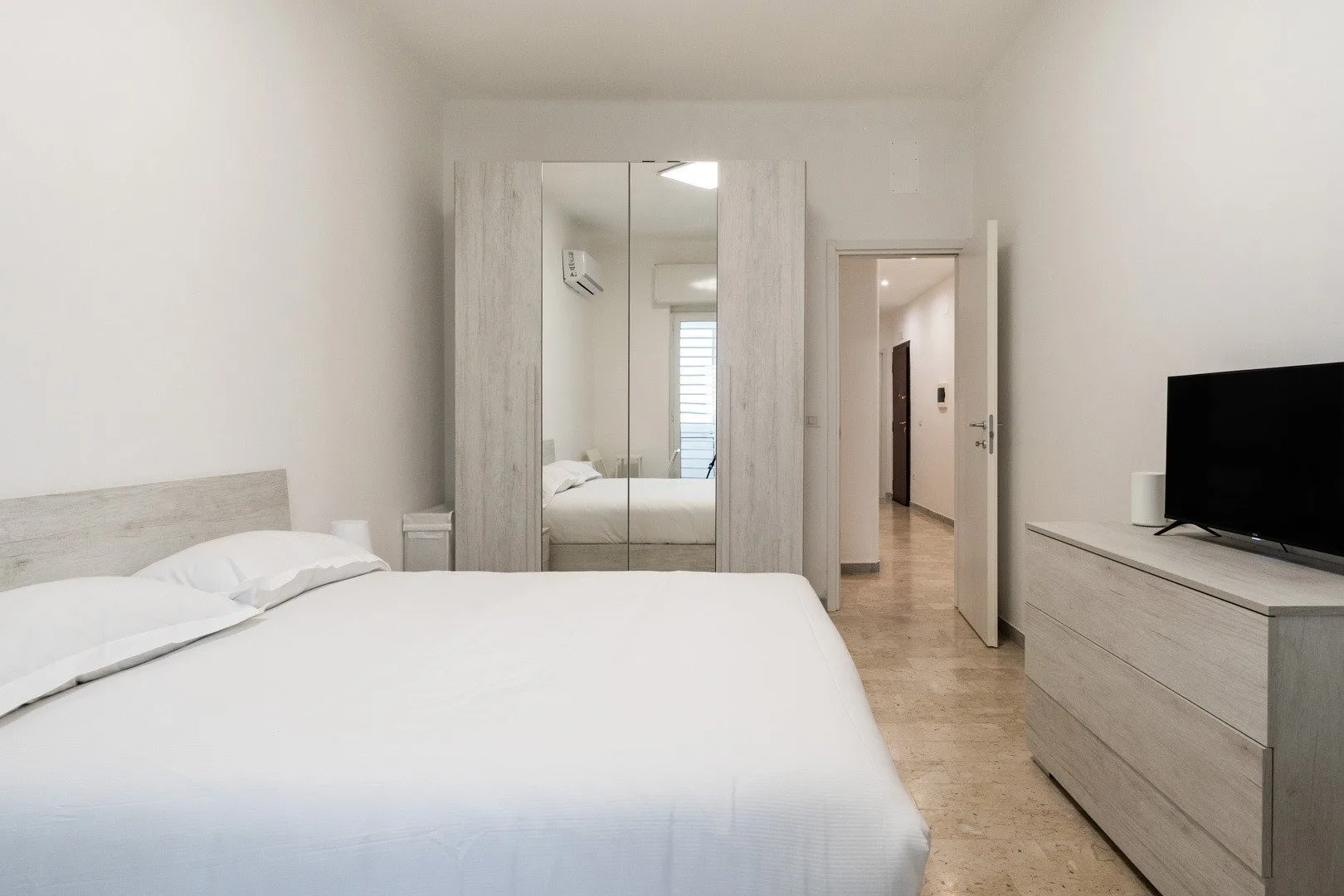 Un dormitorio luminoso con una cama, un armario con espejo y un televisor sobre una cómoda.