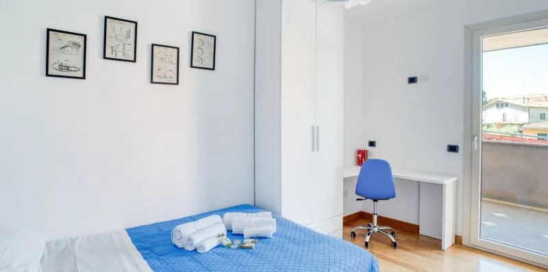 A bedroom with a blue bedspread, folded towels, and a desk beside the balcony door.