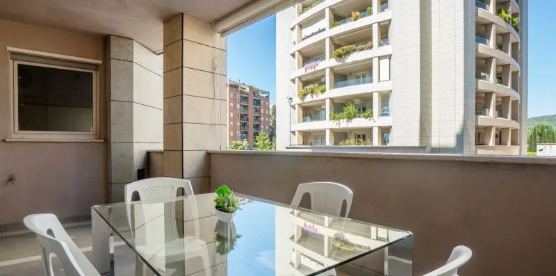 A glass dining table and chairs sit on a covered balcony overlooking nearby buildings.