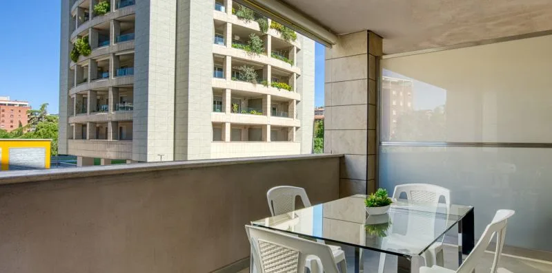A covered balcony with a glass dining table and seating overlooks nearby buildings.