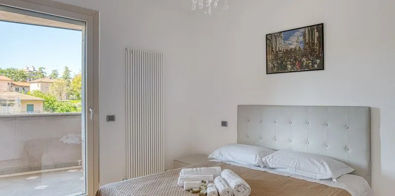 A bedroom with a bed, wall-mounted radiator, and a large window opening onto a balcony.