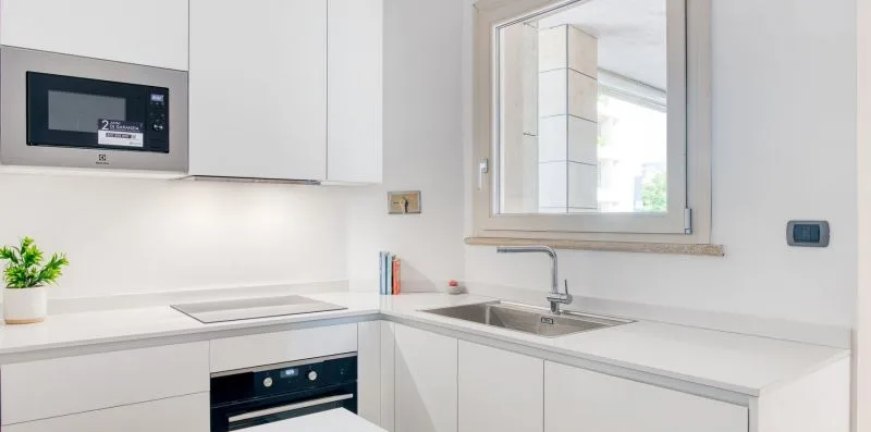A bright white kitchen with a built-in oven, countertop, and sink beneath a window.