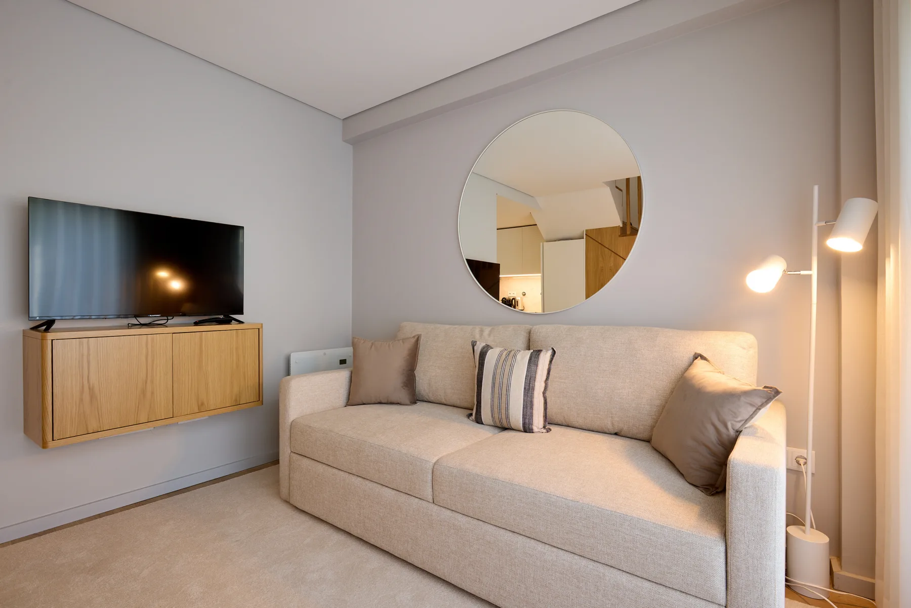 A living room with a light-colored sofa, wall-mounted television, and round mirror.