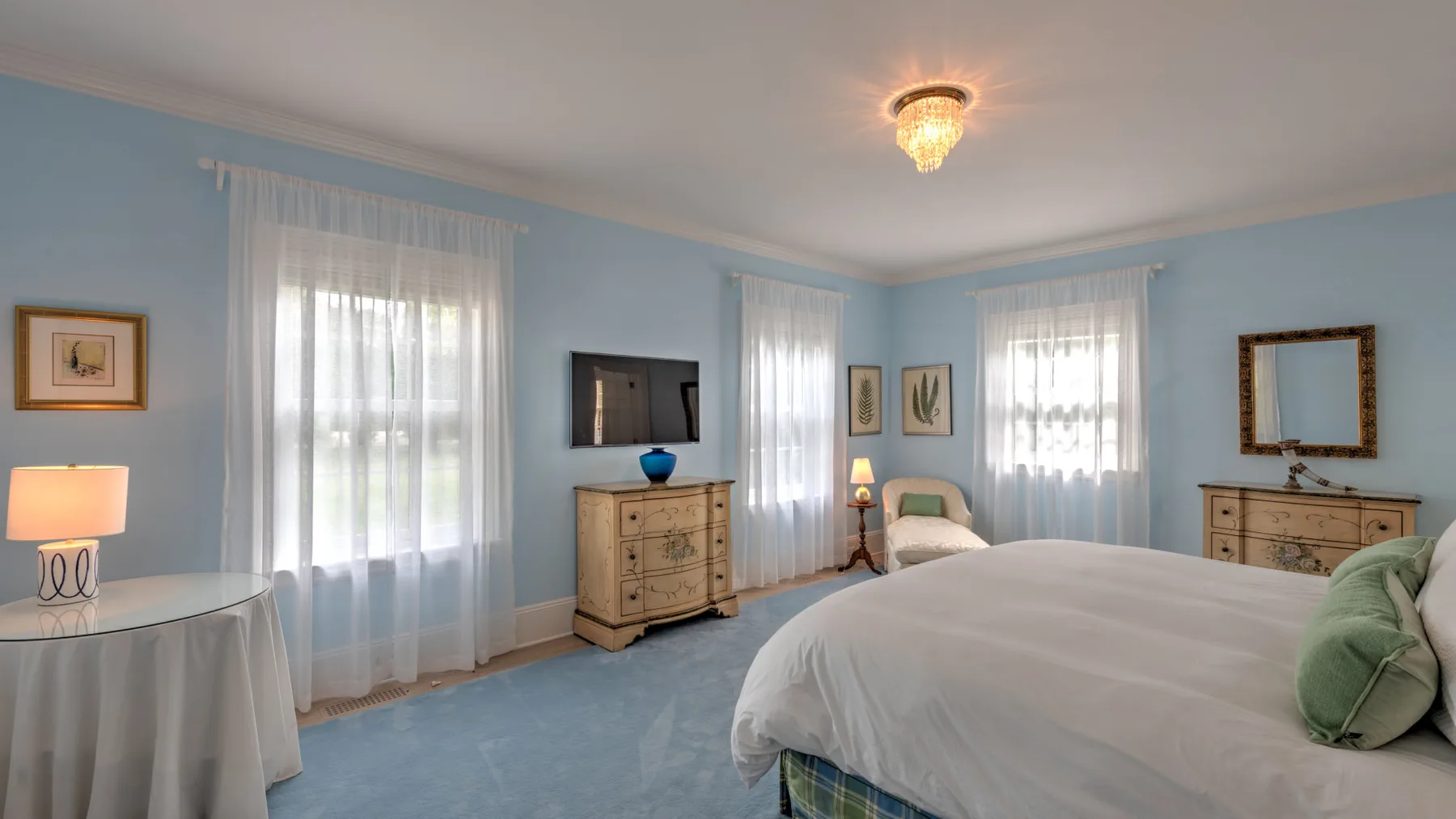 Un dormitorio azul claro con una cama, un asiento junto a la cama y ventanas altas con cortinas.