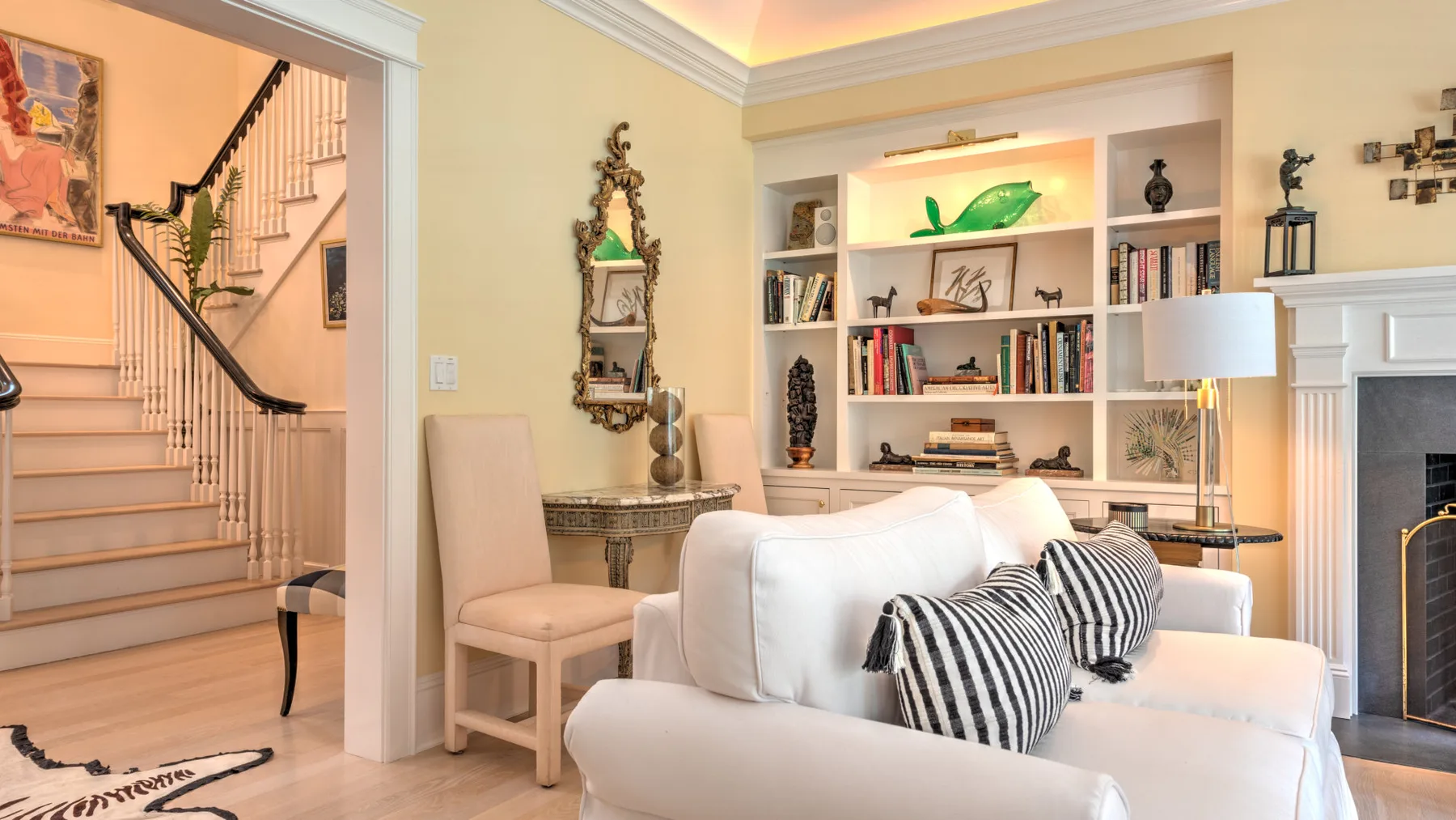 Una sala de estar con asientos blancos, una estantería y una escalera visible a través de una puerta abierta.