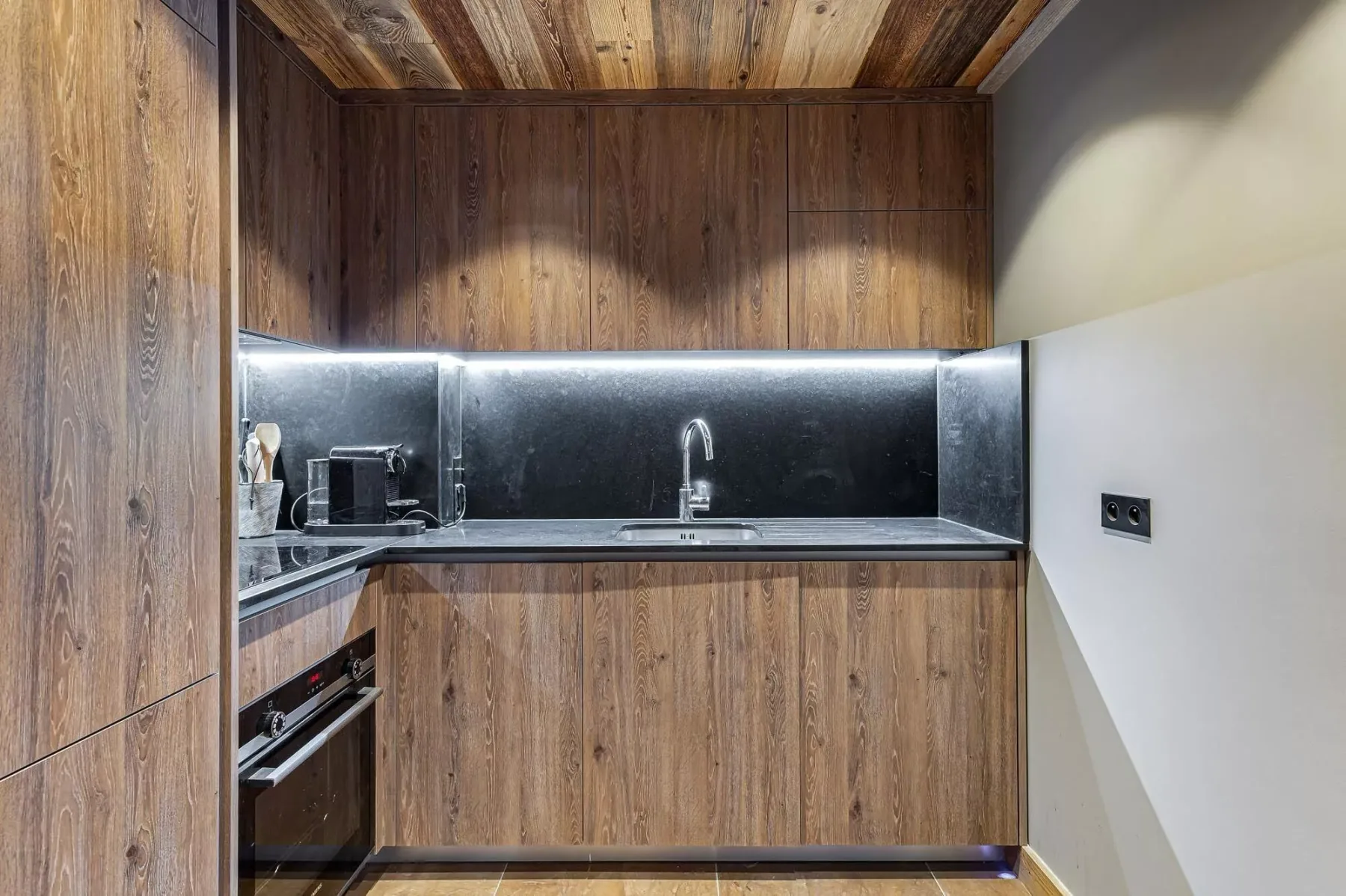 Una cocina compacta con armarios de madera, un salpicadero oscuro y un fregadero.