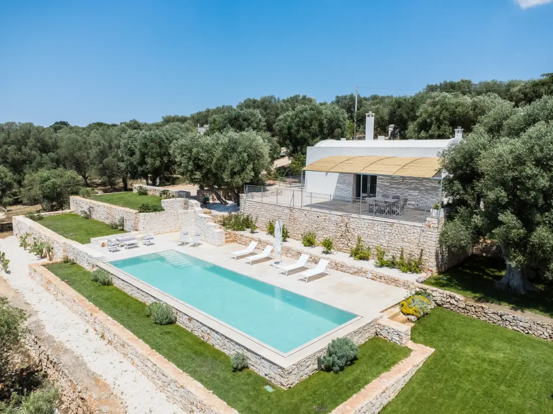 A swimming pool sits beside a stone terrace with loungers and a nearby building.