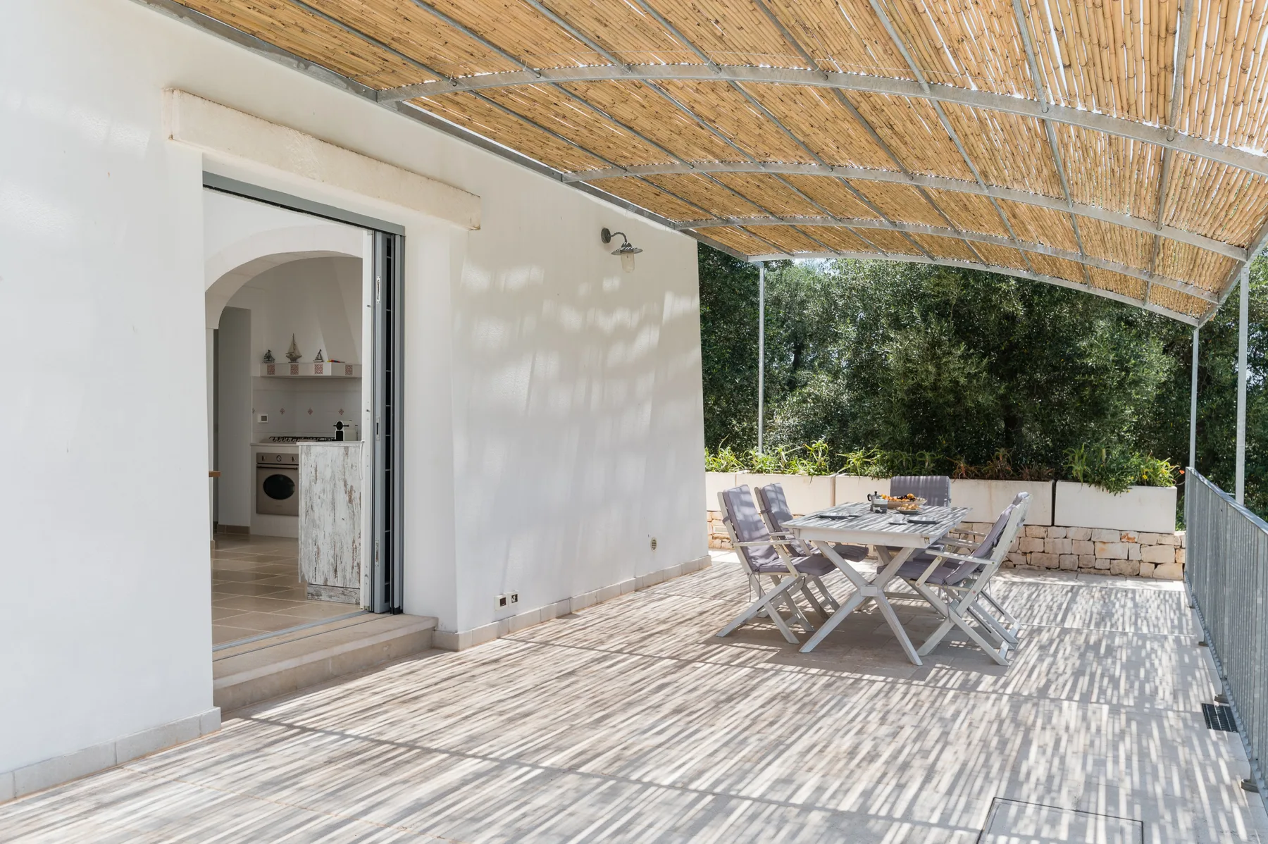 A shaded outdoor dining terrace with a table and seating beside an open kitchen.