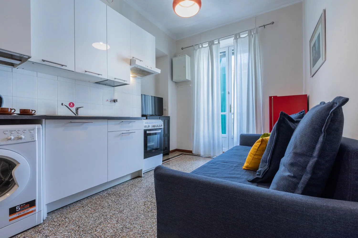 Una cocina compacta y sala de estar con un sofá junto a una ventana luminosa con cortinas.