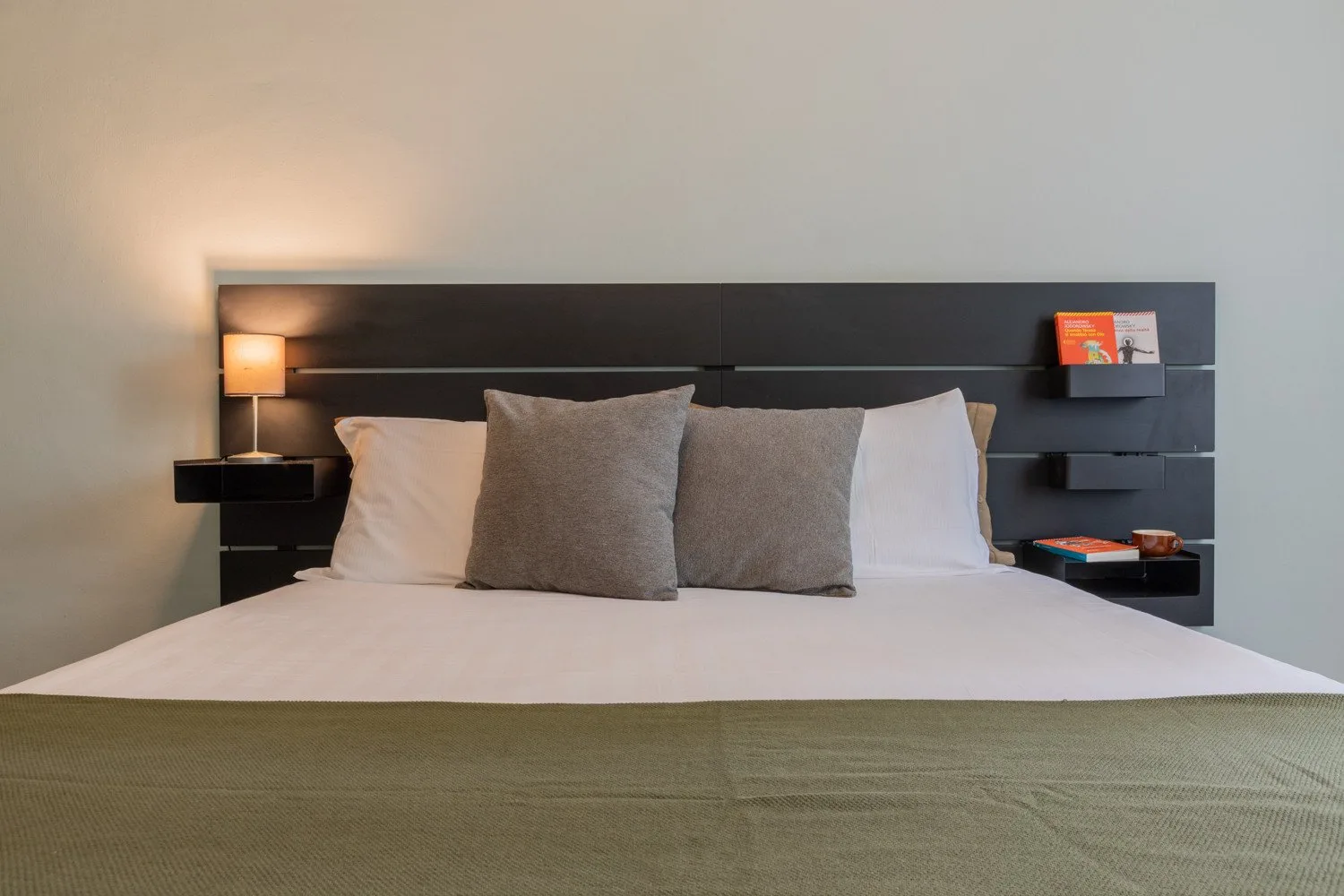 Una cama bien arreglada con ropa de cama blanca y almohadas grises frente a un cabecero oscuro.