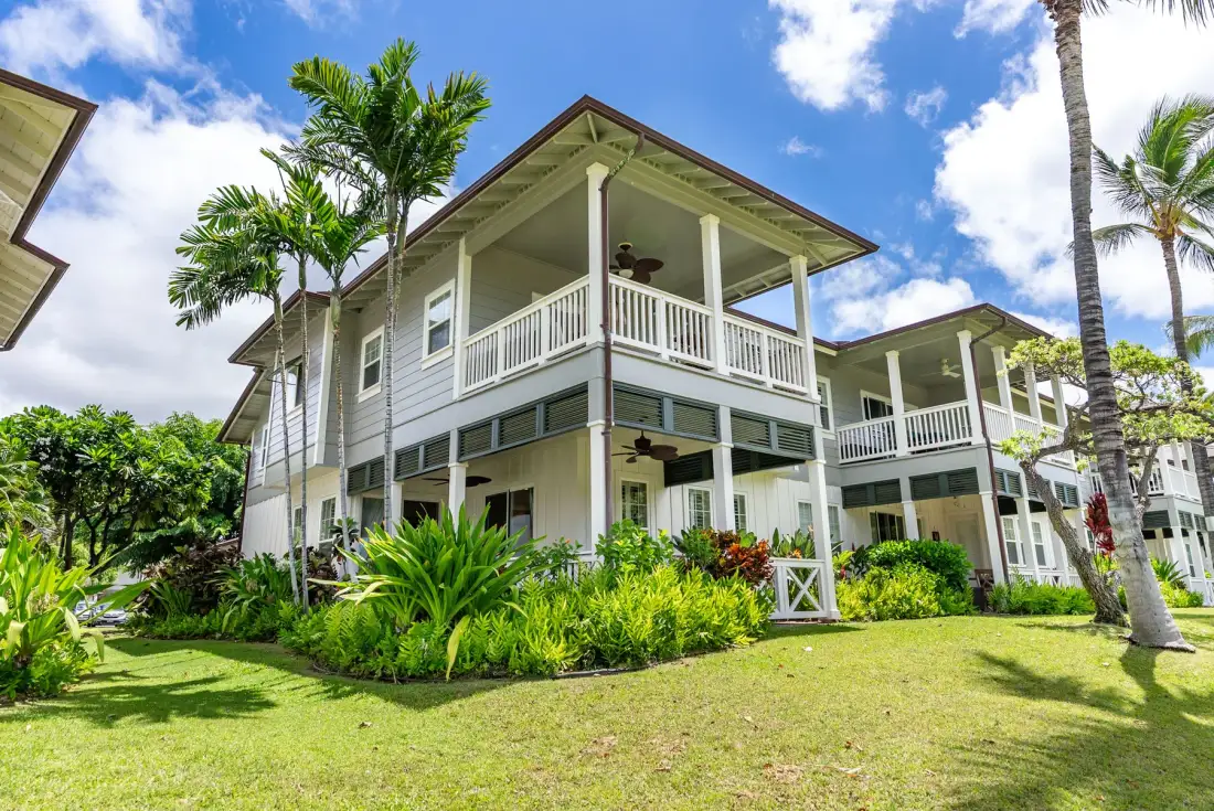 A large white two-story house with wraparound balconies sits among tropical plants.