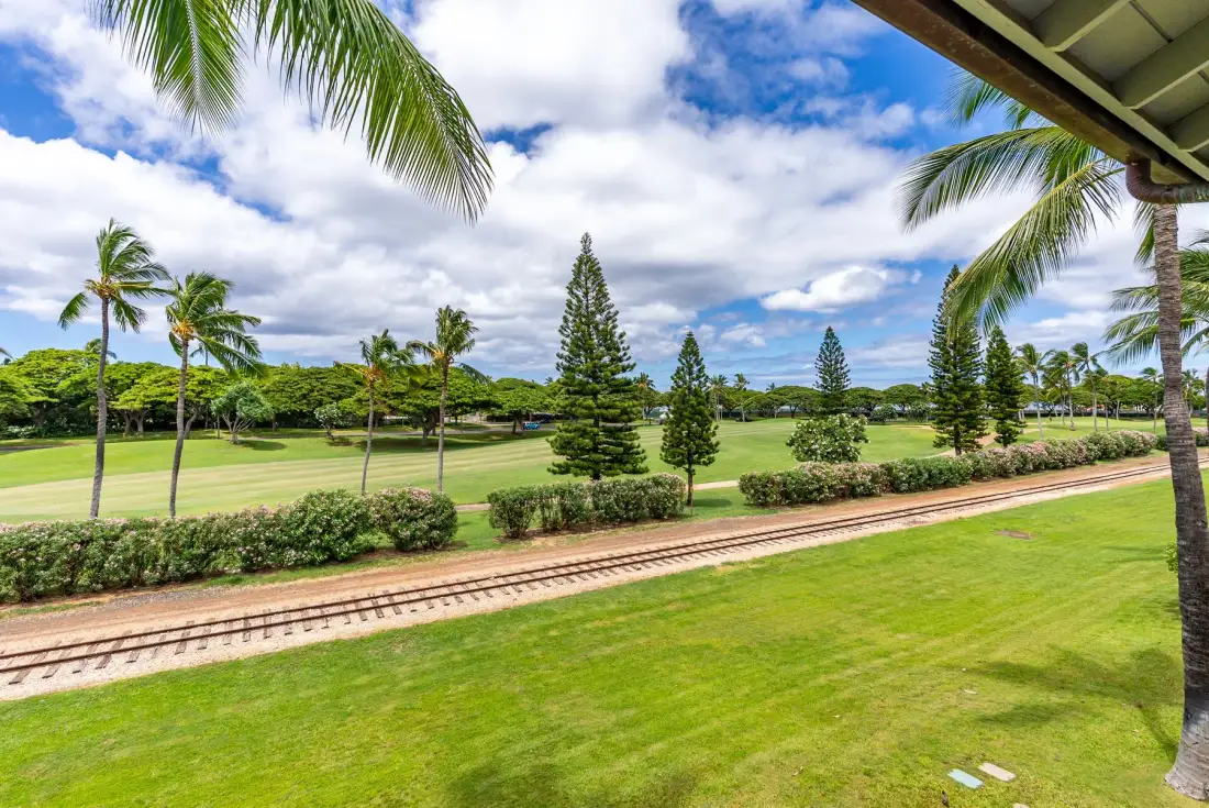 A landscaped green space with palm trees, shrubs, and a narrow rail track under a cloudy sky.