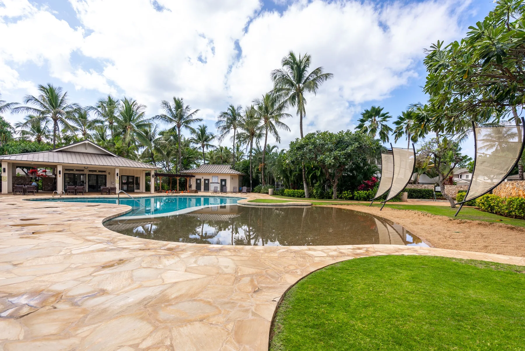 An outdoor pool area with a broad paved deck, lawn, and palm trees.