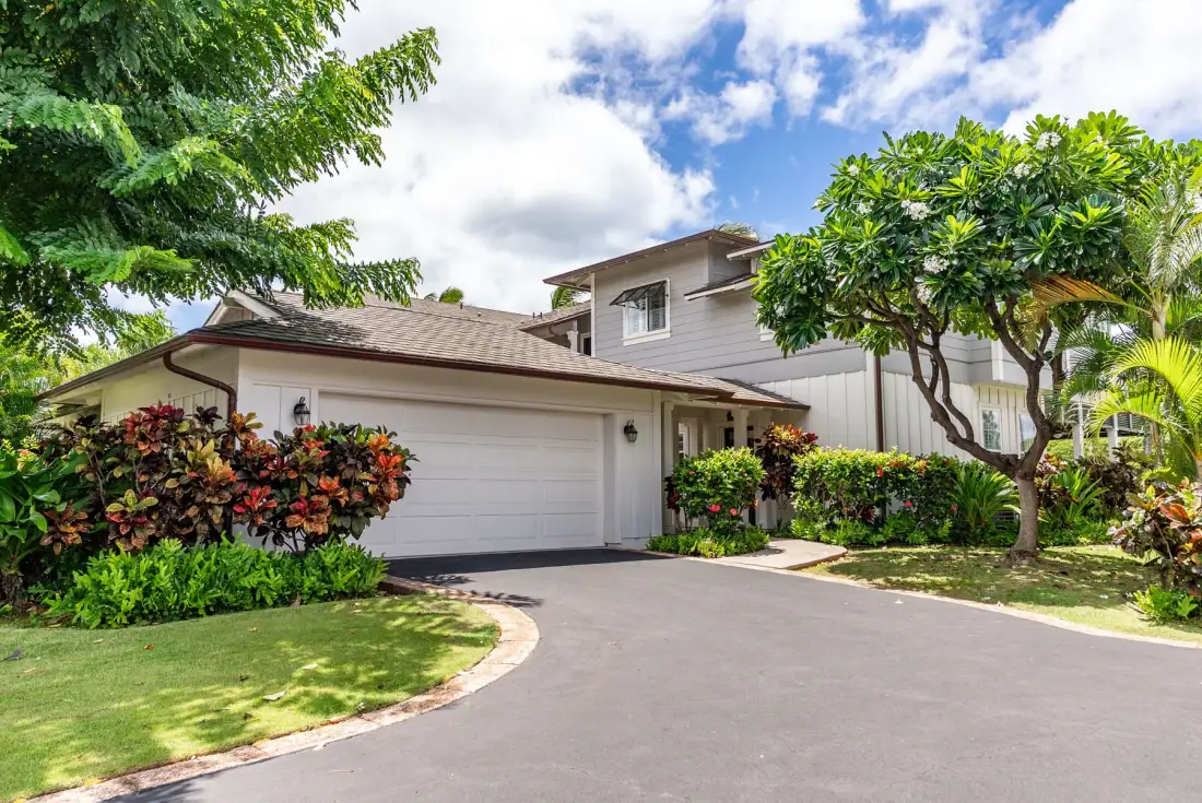 A house with an attached garage sits beside a curved driveway and landscaped greenery.