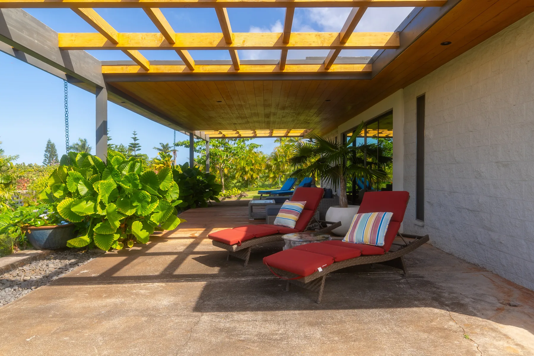 Un patio exterior cubierto tiene asientos acolchados junto a grandes plantas frondosas.
