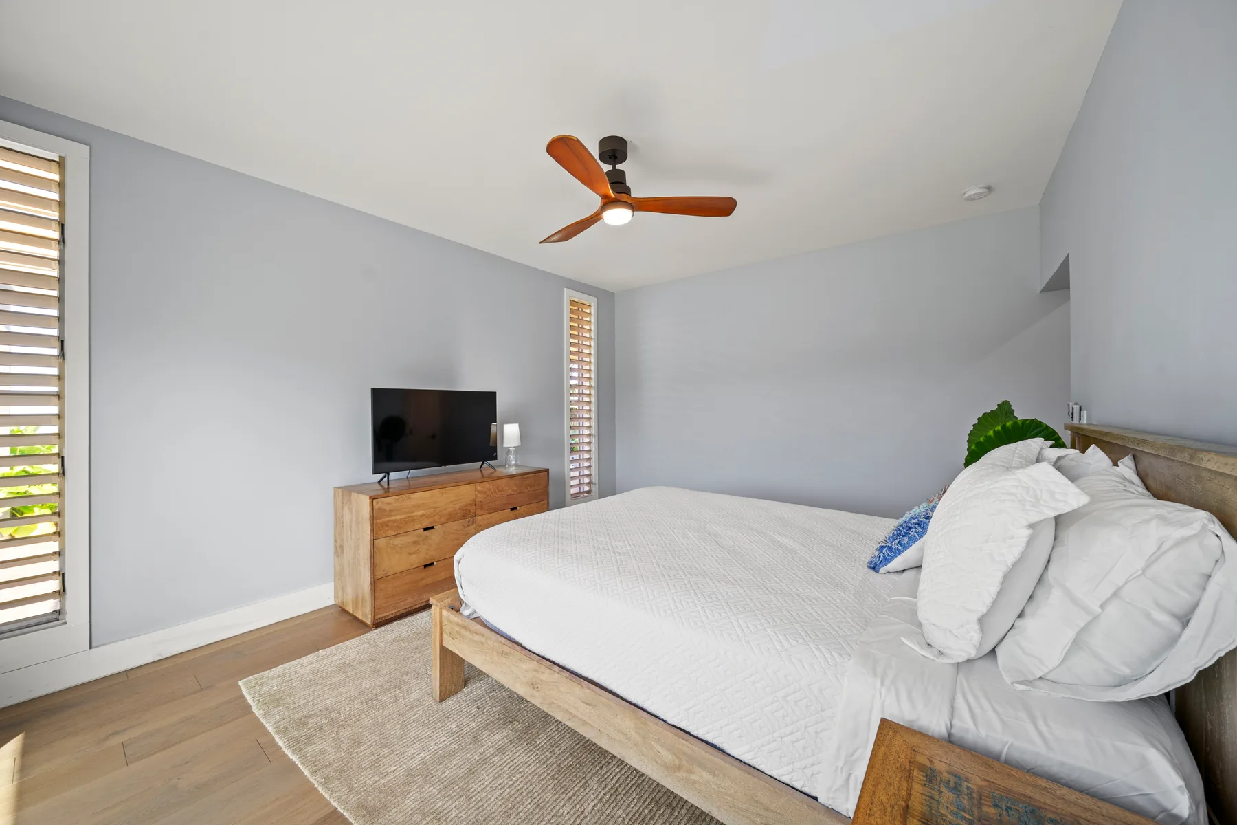 Un dormitorio luminoso con una cama, un televisor sobre una cómoda y un ventilador de techo.